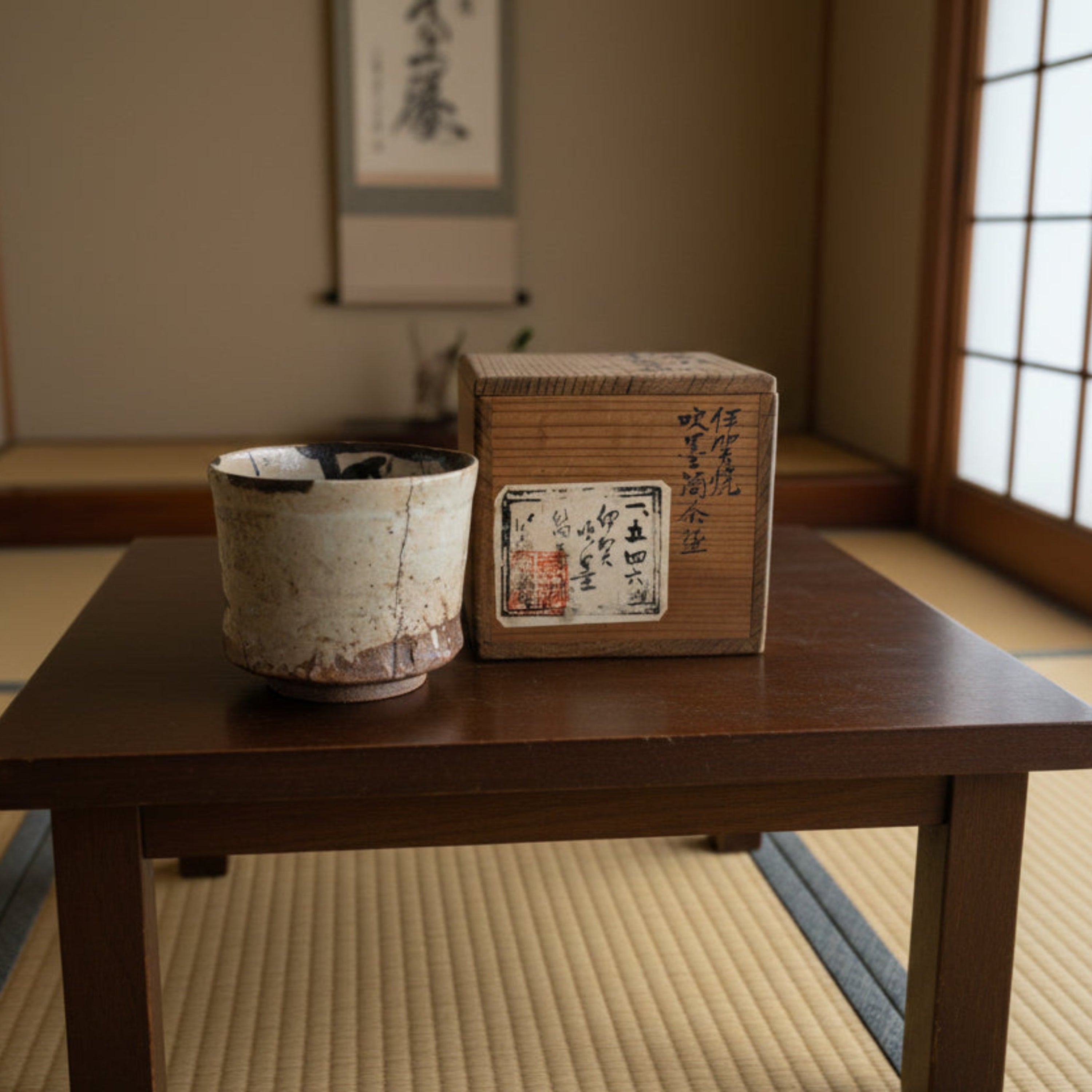 A handmade Japanese ceramic tea cup with a rustic beige and brown glaze, shown next to its wooden presentation box with black calligraphy and a vintage label.